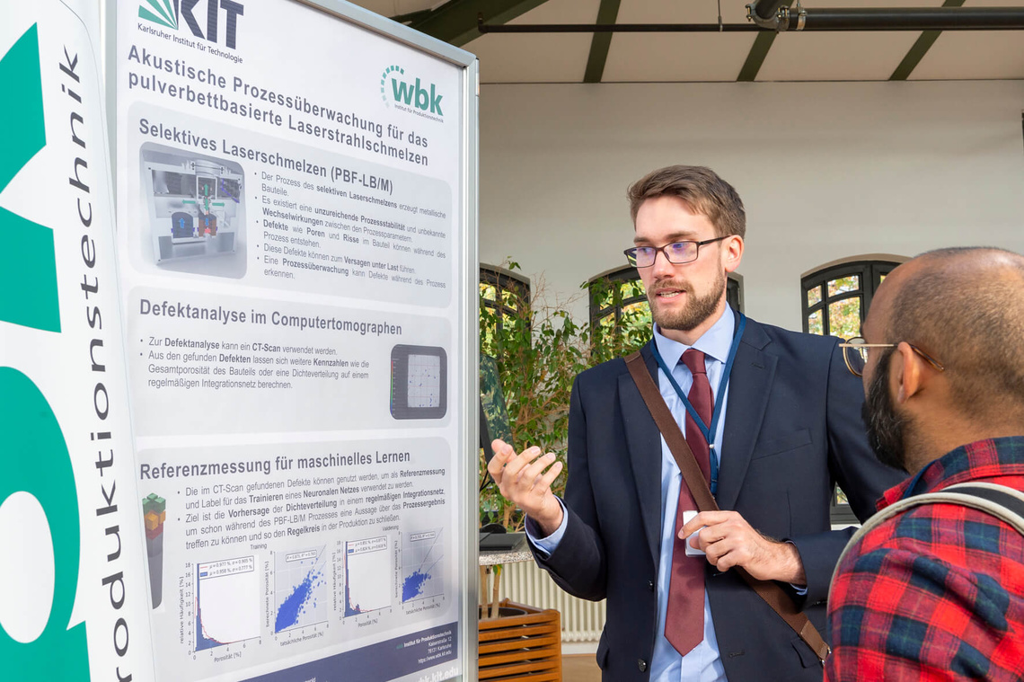 Neben einem Poster "Akustische Prozessüberwachung für das pulverbettbasierte Laserstrahlschmelzen" des Karlsruher Instituts für Technologie stehen zwei Männer und unterhalten sich.